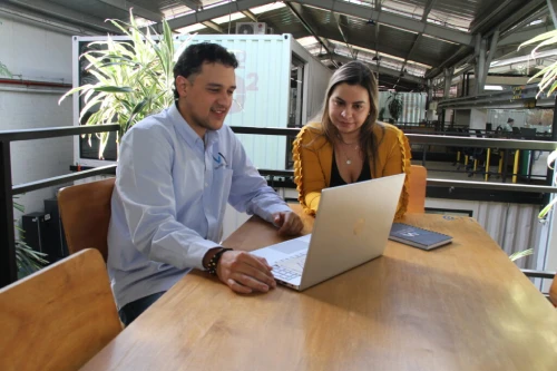 hombre de camisa azul y mujer de chaleco amarillo sentados frente a una mesa en la oficina de vm global group mirando un portátil