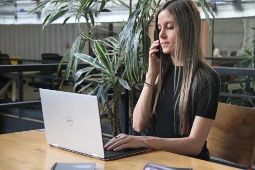 mujer con camisa negra del equipo de trabajo de vm global group, atiende una llamada en su celular mientras digita en su portátil