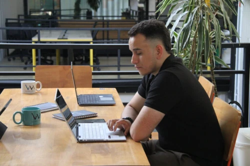 hombre de camiseta negra sentado frente a una mesa de madera y digitando en su portátil, dentro de la oficina de vm global group